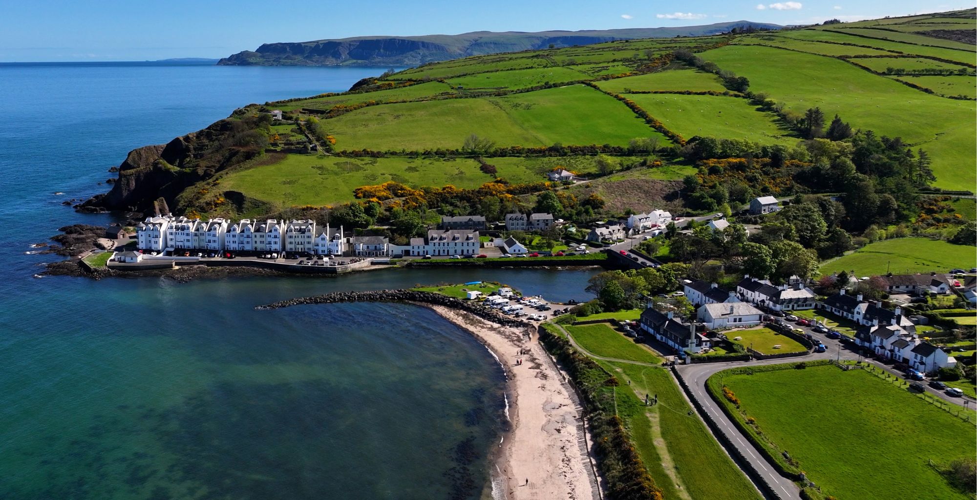 Coastal village with white houses along a sandy beach and clear blue water, green fields on rolling hills behind.